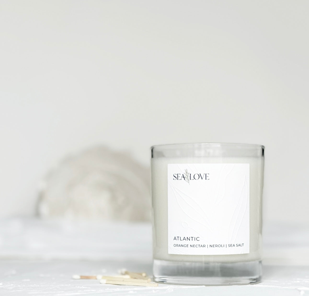 A scented candle in a clear glass jar with a label that reads "sea love, atlantic orange nectar neroli sea salt," placed on a neutral background with a white seashell and matches nearby.
