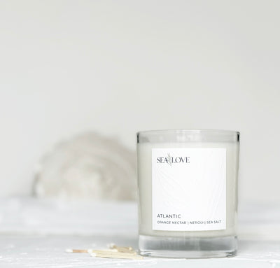 A scented candle in a clear glass jar with a label that reads "sea love, atlantic orange nectar neroli sea salt," placed on a neutral background with a white seashell and matches nearby.