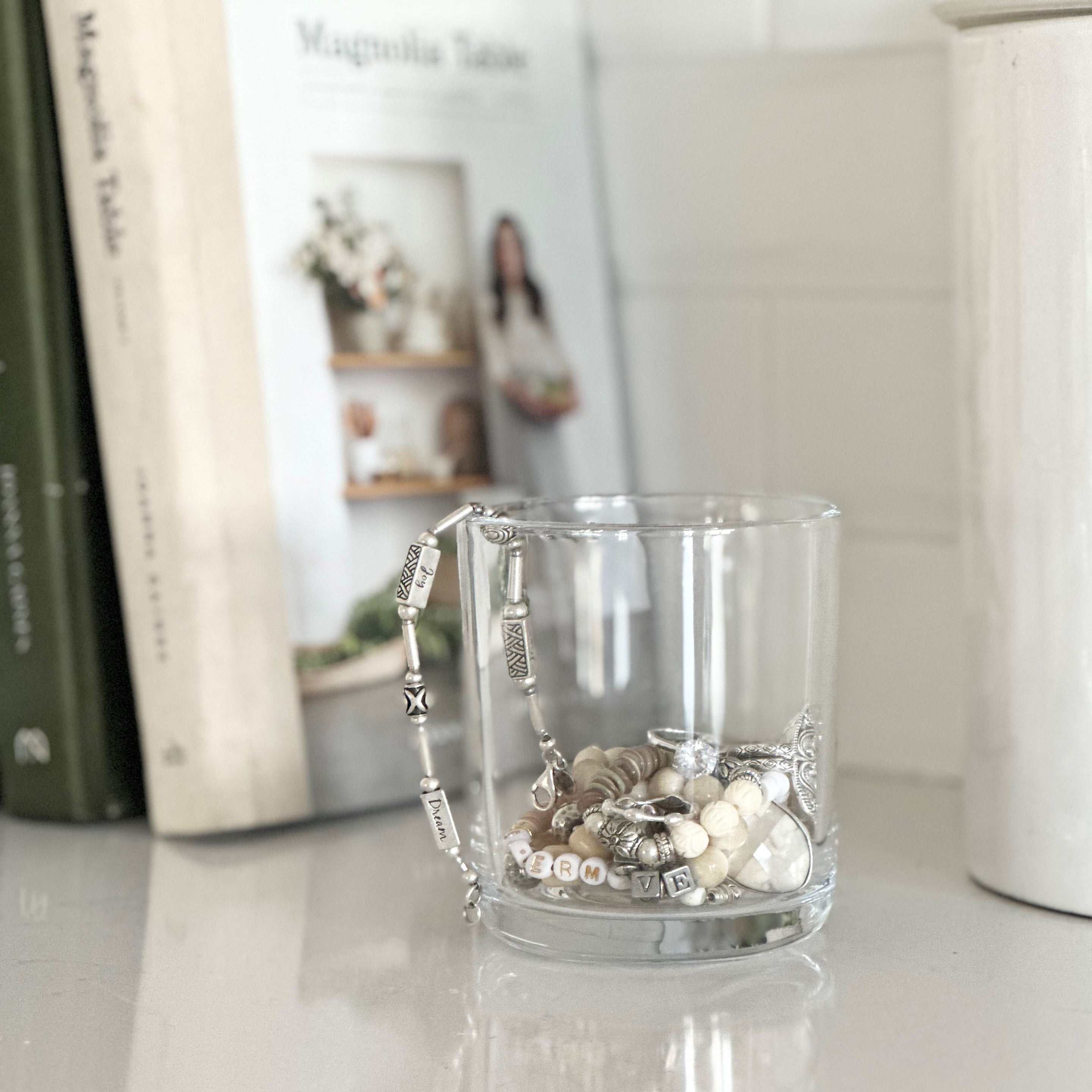 A glass cup overfilled with various charms and beads, sitting on a white shelf next to a row of books, creating a serene and aesthetic vignette.