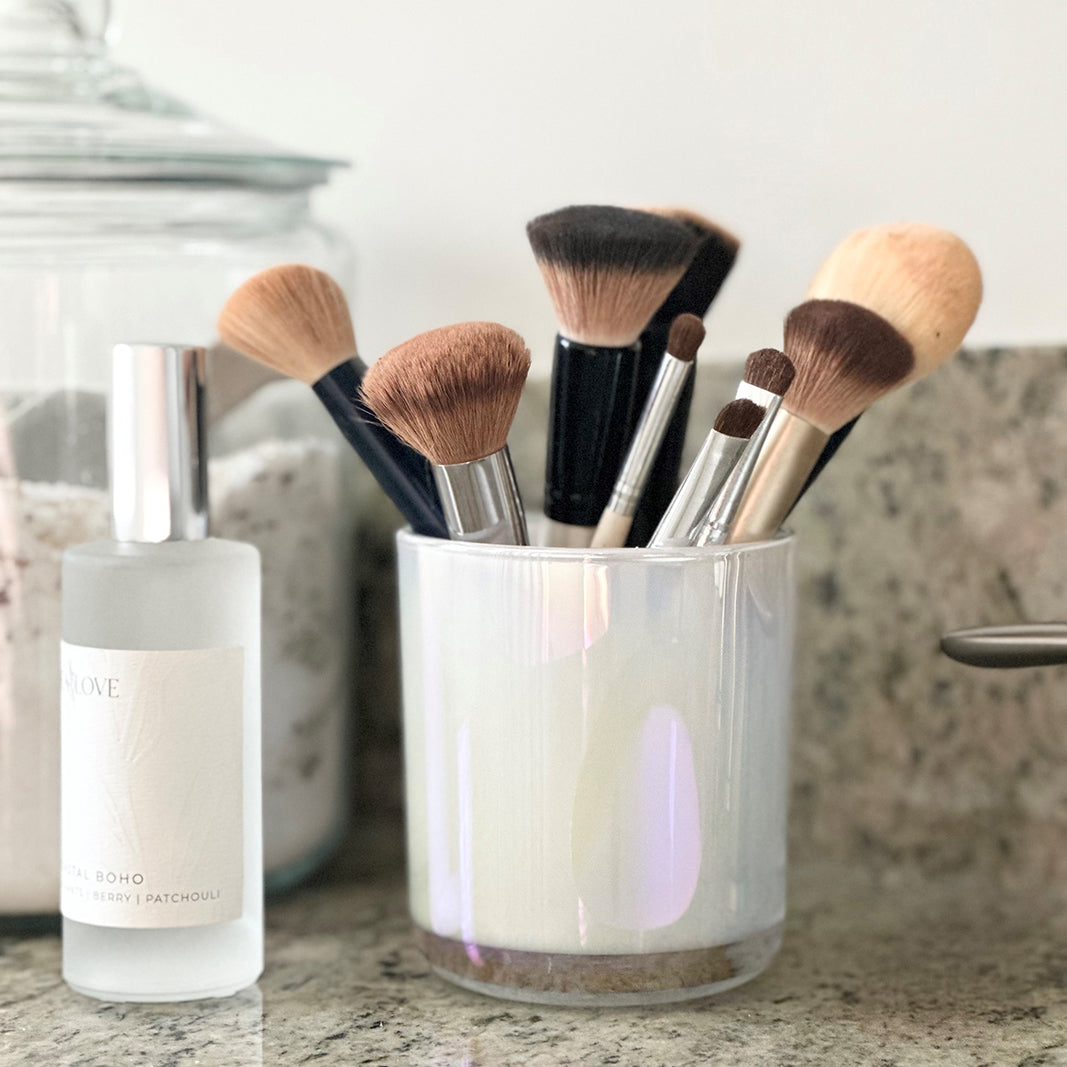 A collection of clean makeup brushes neatly organized in a clear glass holder on a bathroom countertop, next to a bottle of cosmetic product.