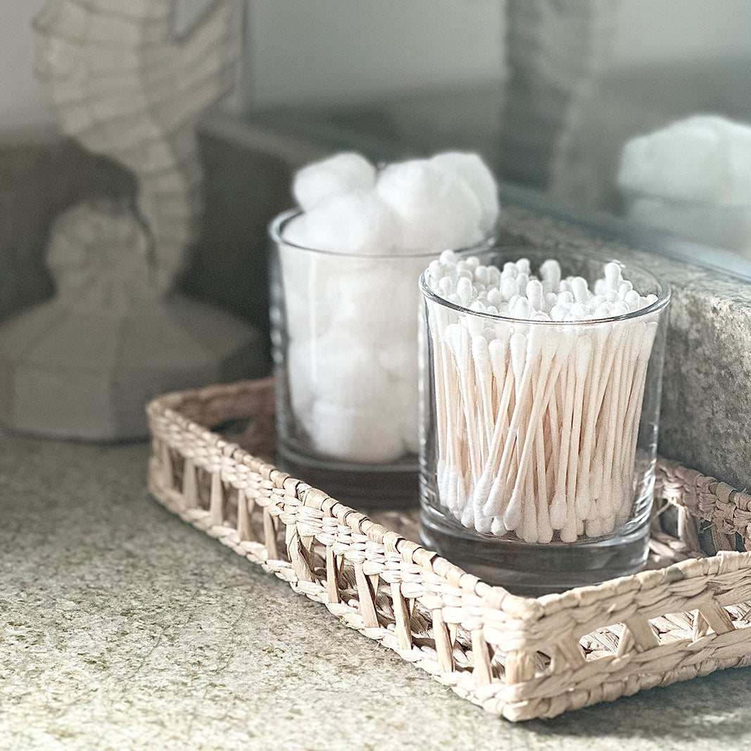 A neatly organized bathroom vanity with cotton balls and cotton swabs stored in clear glass jars on a wicker tray, creating a serene and orderly atmosphere.