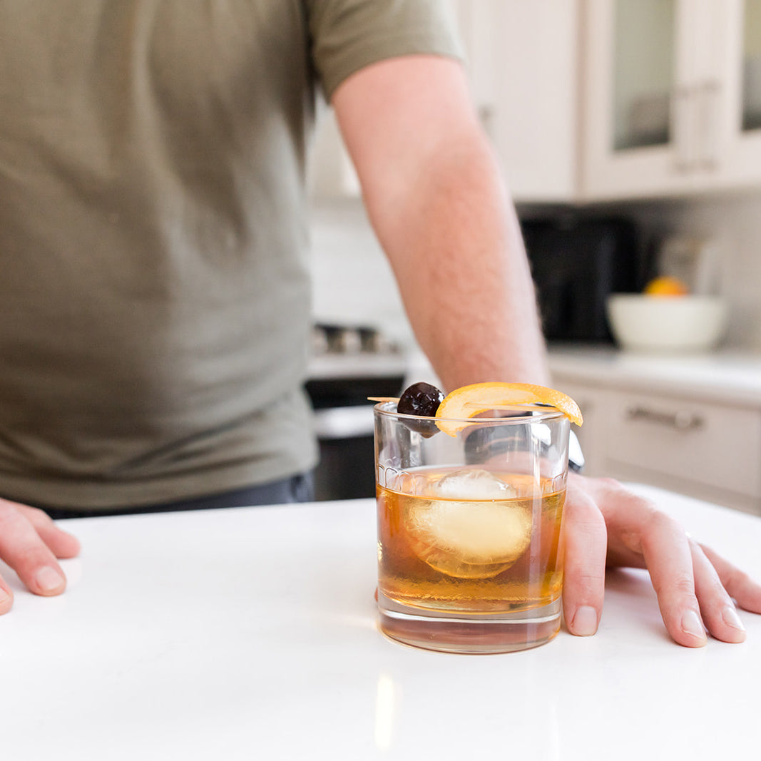 A person standing in a kitchen with a hand on the counter next to a chilled glass of whiskey garnished with a twist of citrus peel and a cherry.