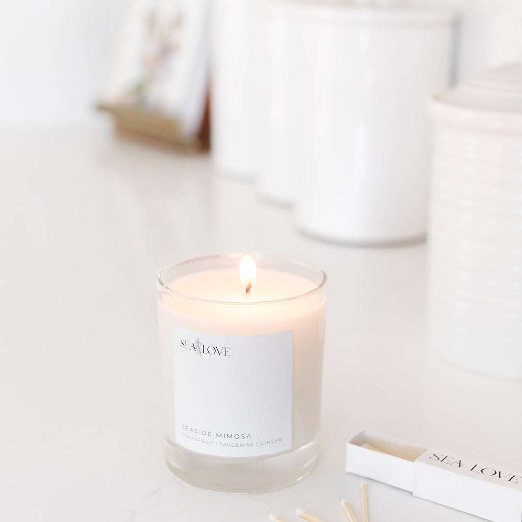A lit scented candle with a label reading "sea love" and "seaside mimosa" placed on a bright, clean surface, accompanied by a matchbox, creating a serene and cozy atmosphere.